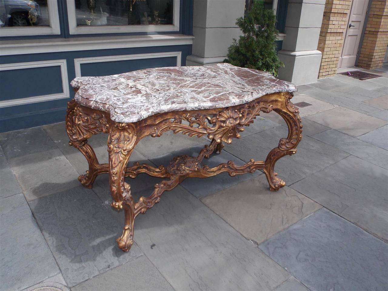French serpentine marble-top gilt center table with floral carved skirts, floral cross stretchers with centered ribbon basket, and terminating on scrolled floral cabriole legs. All original, Mid-18th century.