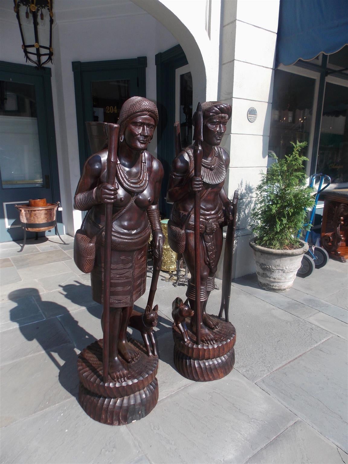 Pair of Igorot tribesmen figural wood carvings depicting hunter and huntress with companion canines, weaponry, and displayed on circular fluted plinths. Northern Luzon , Philippine Islands . Were feared & known as deadly head hunters. Mid 20th