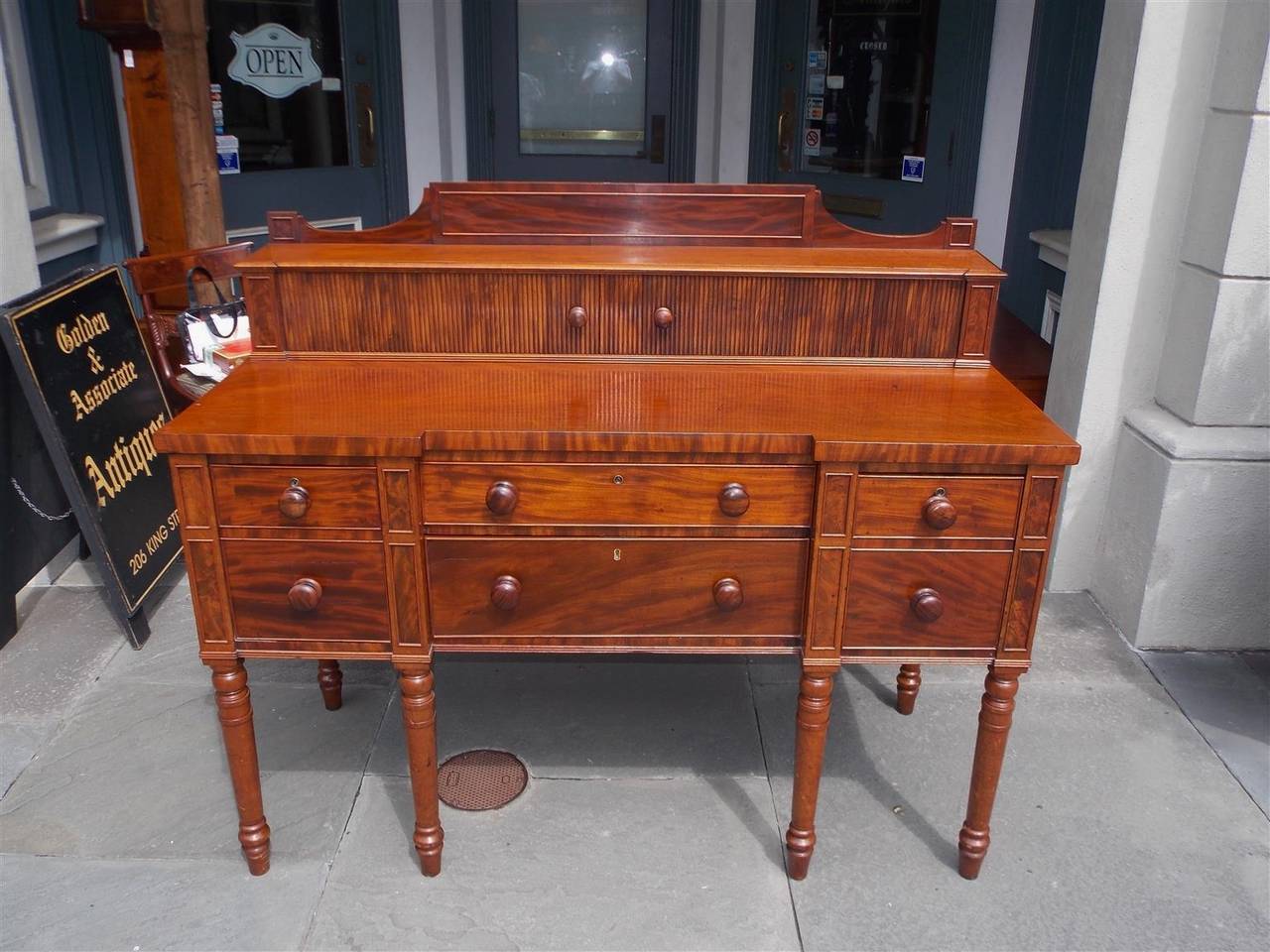 English Regency mahogany sideboard with scalloped tambour top back splash, ribbon mahogany molded edge panels, two over two lower case center drawers and flanking cellarette drawers with the original wooden knobs and terminating on the original