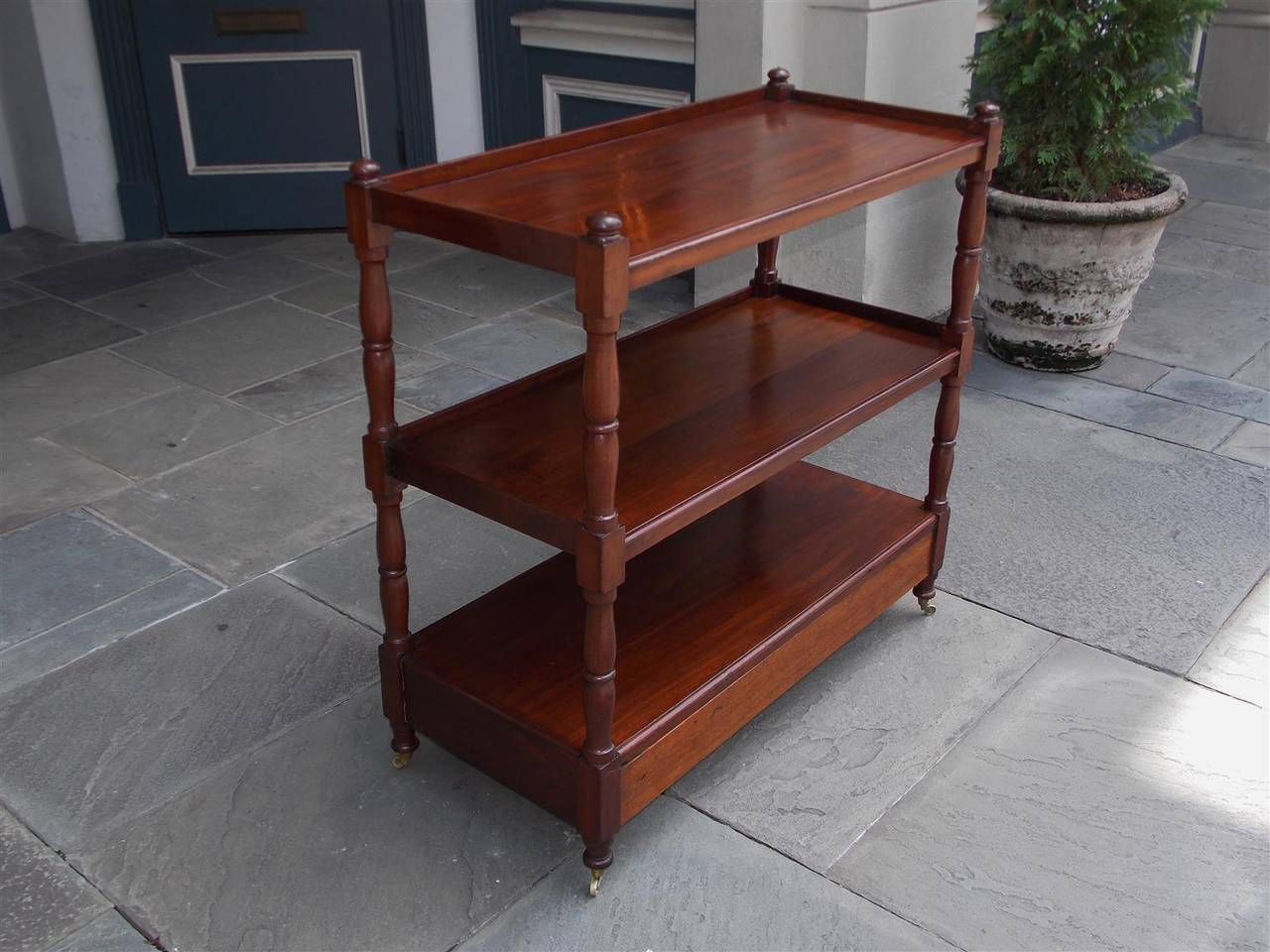 English mahogany three-tiered trolley with squared turned ringed bulbous post, ball finials, single lower drawer and terminating on original brass casters. Trolley is finished on all sides, Early 19th century.