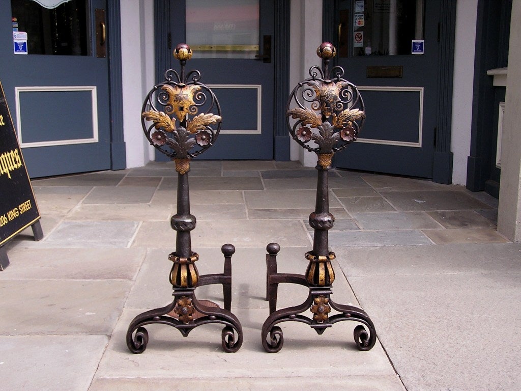 Pair of Italian gilt figural mask wrought iron and poly chromed medallion andirons with flanking gilt foliage and painted ball finials, decorative scrolled gilt foliage iron work, central tapered gilt fluted plinths, flanking ball finial hammered