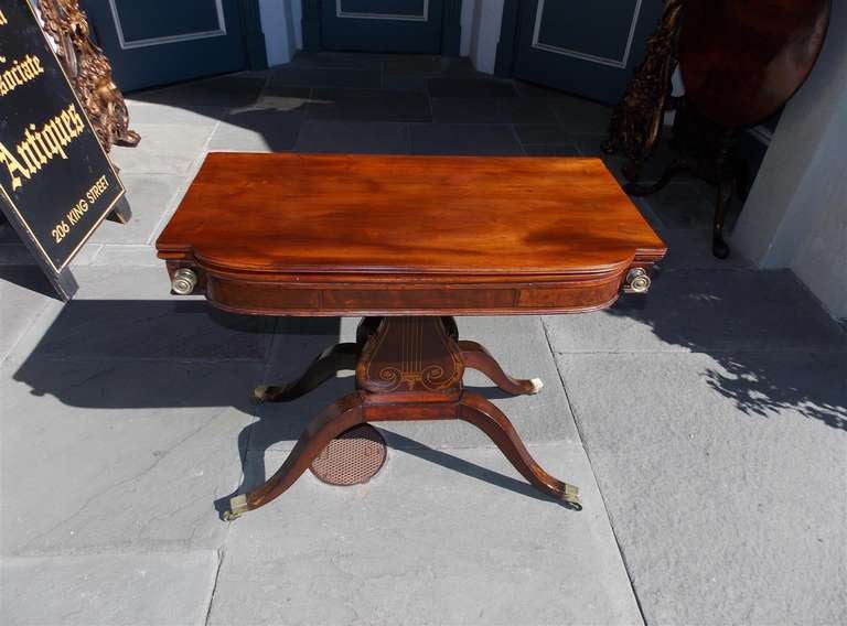 American mahogany flip top game table with hand stenciled centered lyre pedestal, resting on saber legs with stenciled acanthus motif and original brass casters.