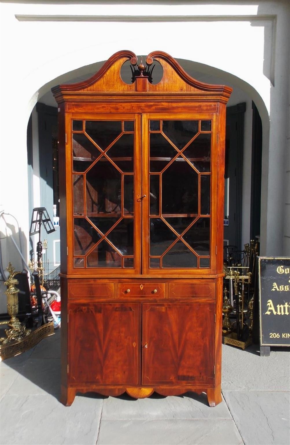 American Mahogany elegant corner cupboard with a centered urn scrolled inlaid pediment, original upper flanking glass hinged doors, fitted interior butterfly shelving, single centered drawer, original brass pulls, lower flanking blind hinged doors ,