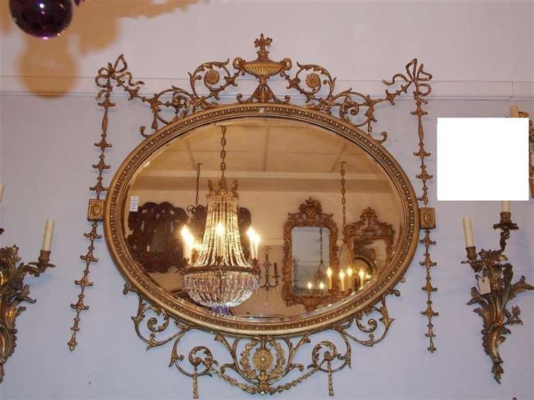 English Adam style gilt carved wood and gesso oval wall mirror with centered decorative urn, scrolled acanthus foliage medallions, flanking corner ribbons accompanied by graduated foliate bell flowers.  Mirror retains the original oval beveled glass.