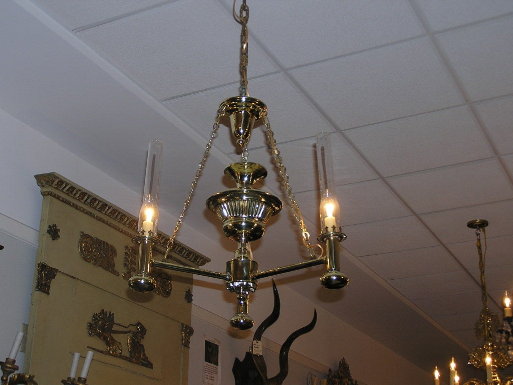 American three arm brass argand chandelier with ribbed centered urn, glass chimneys, original chain & canopy, and terminating on floral finial. Originally oil and has been electrified.