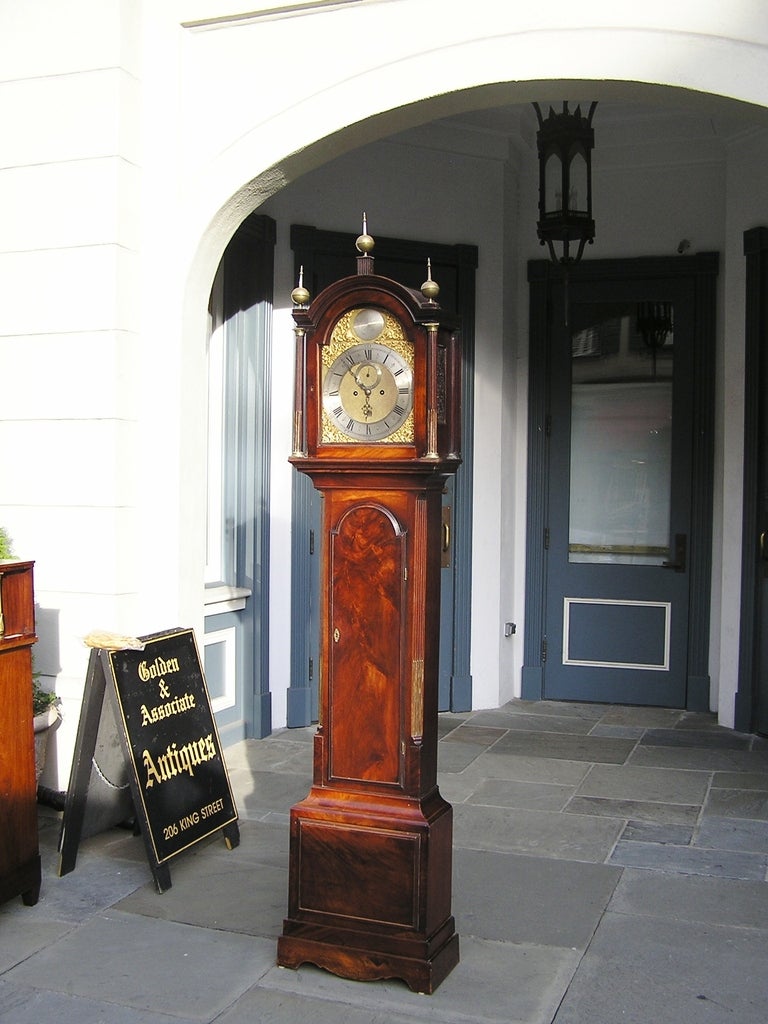 English Mahogany tall case clock with brass finials, arched bonnet with brass fluted columns, gilt ormolu and chased polished steel face, flanking reeded quarter columns, and terminating on a molded squared base with serpentine skirt. Late 18th