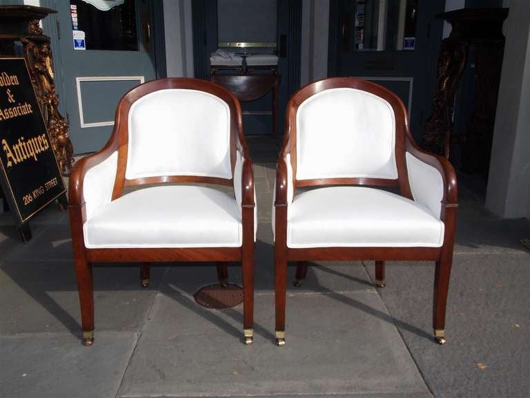 Pair of French mahogany bergere chairs with scrolled back and arms, upholstered in white muslin, terminating on tapered squared legs with original brass casters.  Circa 1820