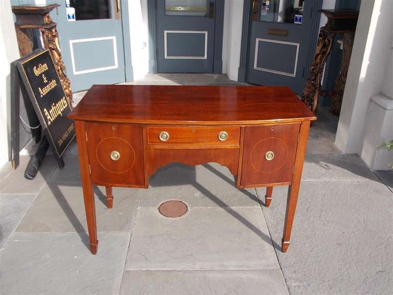 English mahogany bowfront brandy board with centered one drawer, flanked by matching side cabinets, circular satinwood inlay, and terminating on tapered spayed legs.  Circa 1810