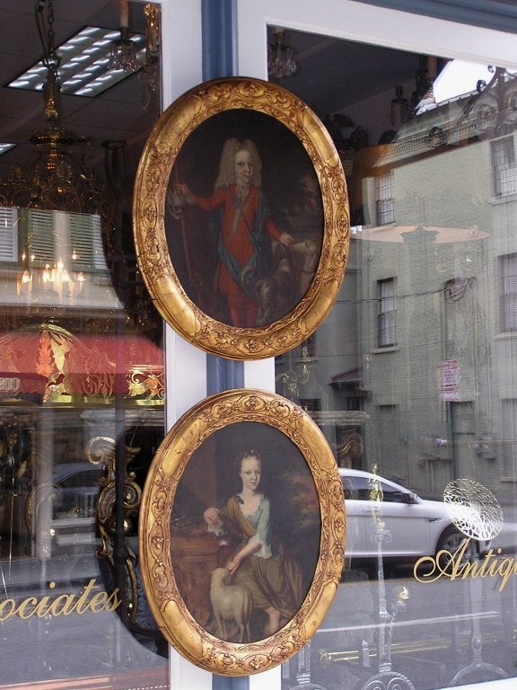 Pair of English oval oil on canvas portraits of young male posing with hunting canine companions and young female sitting in garden posing with flowers and lamb. Paintings are in original gold gilt carved oval frames.