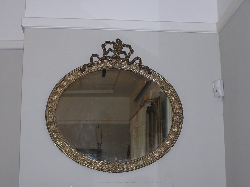 French Silver Gilt Tin Oval Wall Mirror with Central Ribbon Cartouche and Flanking Foliage Pinwheel Medallions, Mid 19th Century.  Mirror retains the original glass and rear backing. 