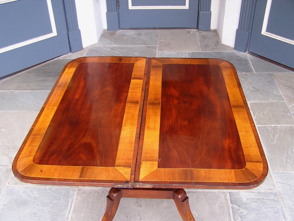 English mahogany hinged game table with book matched cross banded satinwood inlays, ebony sting inlays, turned ringed pedestal columns, and resting on the original saber legs with foliage brass casters. Early 19th Century