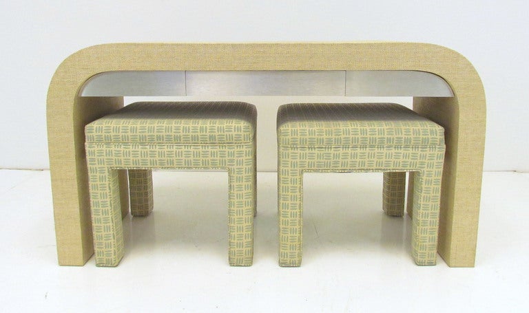 Waterfall form console with grass cloth covering and brushed aluminum apron with drawer, with pair of upholstered stools, ca. late 1970s.   Console is double sided so can float in a room.  In the manner of Karl Springer.

The console table is