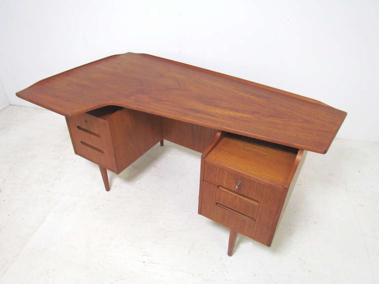 Danish teak desk featuring an asymmetrical top with raised lip edges, in the manner of Arne Vodder or Peter Lovig Nielsen.    Three drawers at right, one large deep drawer at left, the included key functions in both locks.  Reverse side features