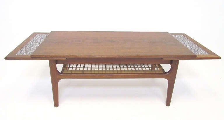 Teak coffee table with two expandable pull-out self storing leaves and cane under shelf, by Trioh, Denmark.   The leaves feature laminate tops with a modernist print design, especially good for your hot and cold serving needs.

41.25