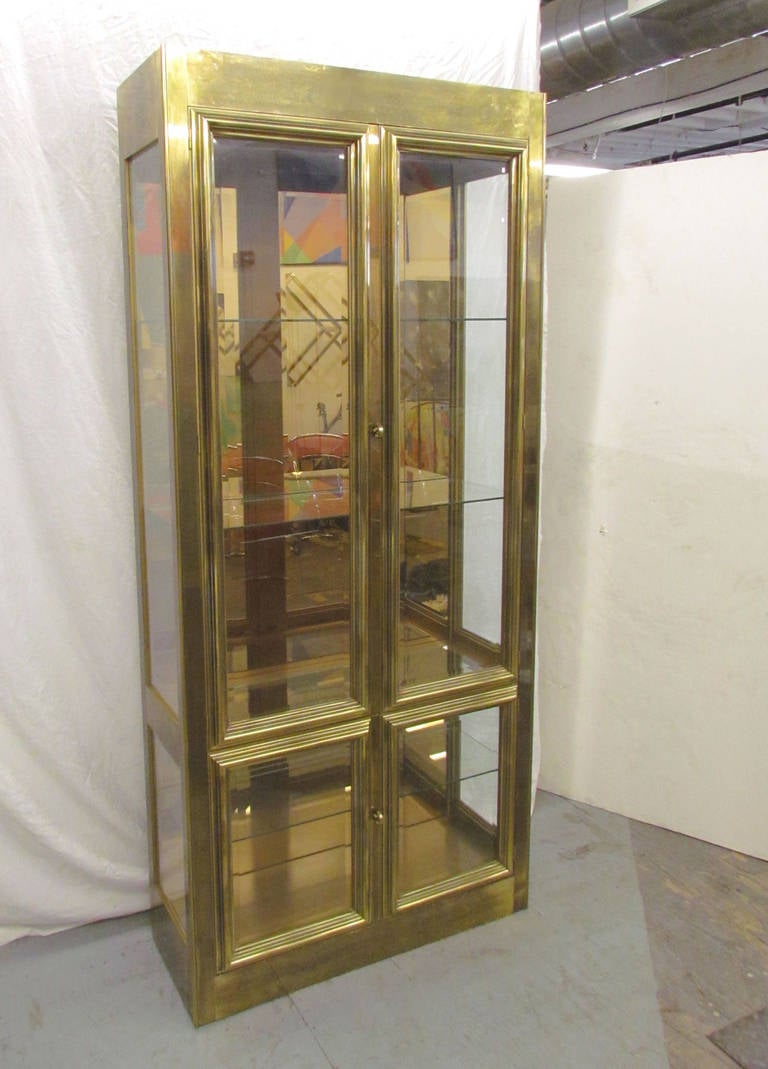 Elegant brass-clad vitrine by Mastercraft, circa 1970s. Two sets of double doors with solid brass hardware, mirrored back, glass shelves with inset ridge for displaying plates or other flat items, recessed interior spot lights. Retains Mastercraft