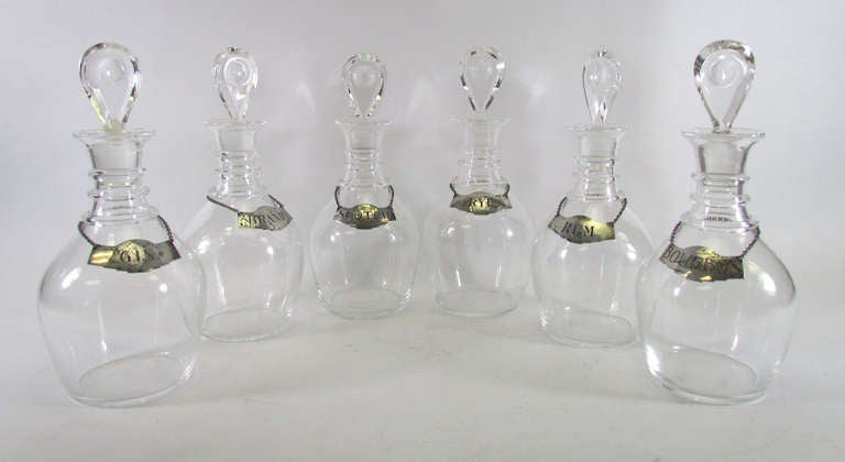 Set of six glass decanters by Steuben Glass, adorned with sterling liquor tags, ca. late 1940s -- early 1950s.   Labels include Brandy, Rum, Scotch, Rye, Gin, and Bourbon.   Each decanter signed Steuben, and each decanter collar signed with the mark