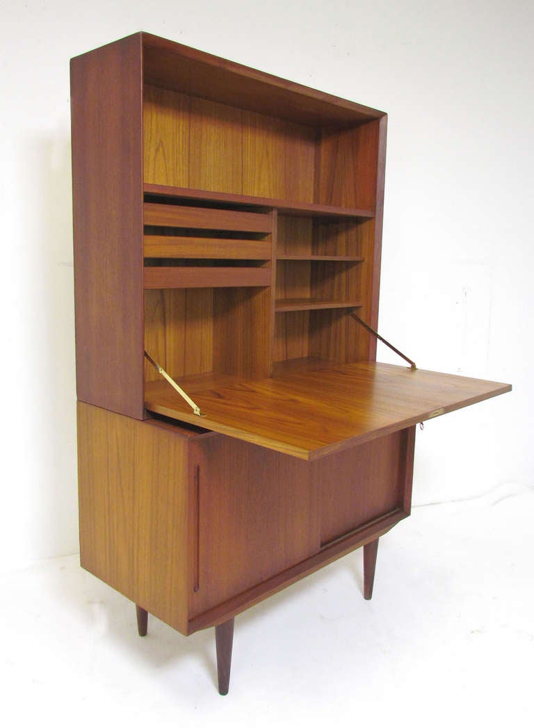 Danish teak two piece secretary consisting of server size media cabinet with pair of sliding doors topped with (removable) upper unit consisting of a generously sized drop down desk with interior storage and stationery drawers.  With original