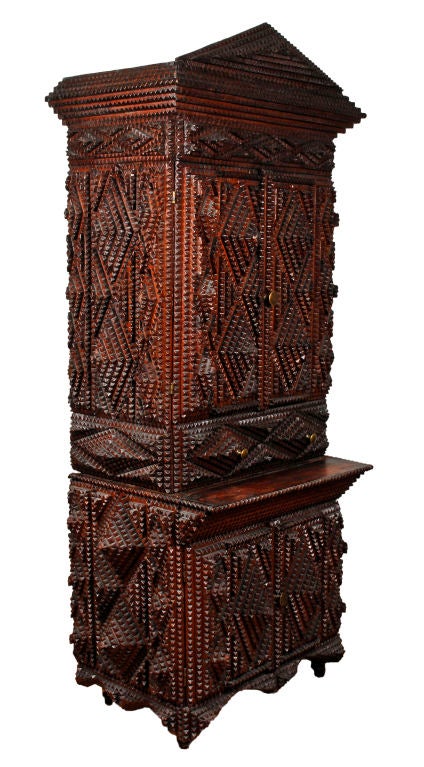 Tramp art scratch built two piece cupboard with an apex top and bold diamond designs. There are cabinet doors on top over a drawer with another set of doors on the bottom. The inside has multiple compartments and retains remnants of its original