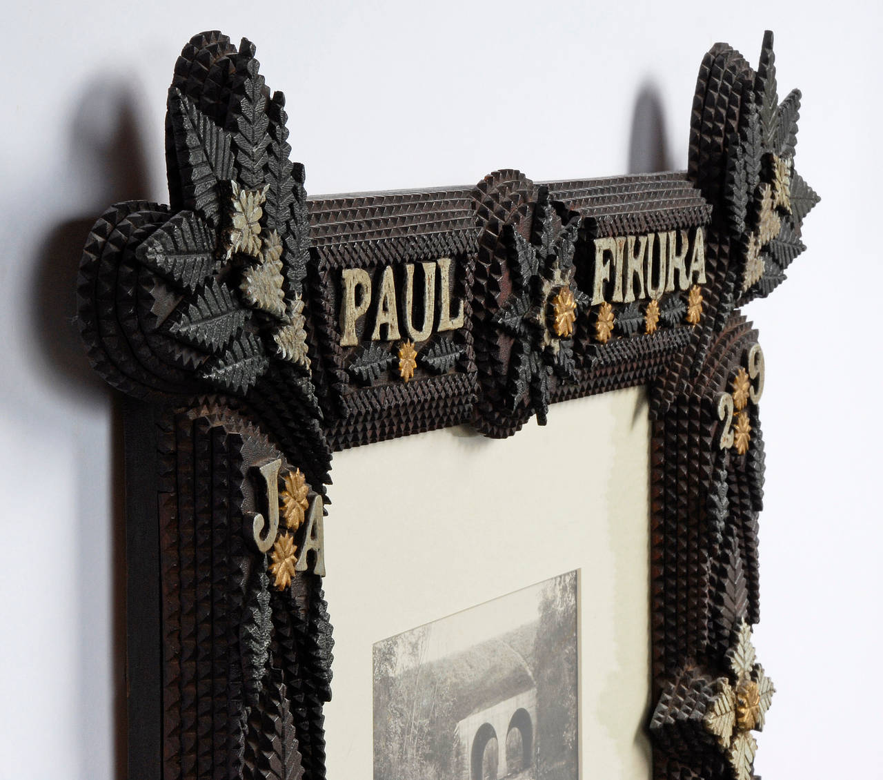 An expressive Tramp Art frame with brightly painted highlights and inscribed, ‘Paul Fikura, JA, 29, 1912′ with carved leaves, hearts, stars, rosettes and trees. A photo of a man fishing in a stream is mounted in the frame. 

Dated JA (January),