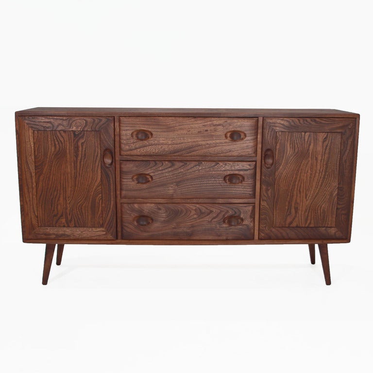 A cabinet made Lucian Ercolani for Ercol, a furniture manufacturer in London. There are three drawers in the middle and two doors on wither side.  On each, there are sculpted handles and handle recesses that are trademark of Ercoliani.  Angled,