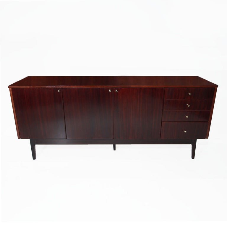 A dark mahogany credenza  with three doors and four drawers opening out. Beautiful wood grain is seen all throughout, and brass knobs make for standout hardware. There are five legs, one of which, in the center, provides for ample support of heavy