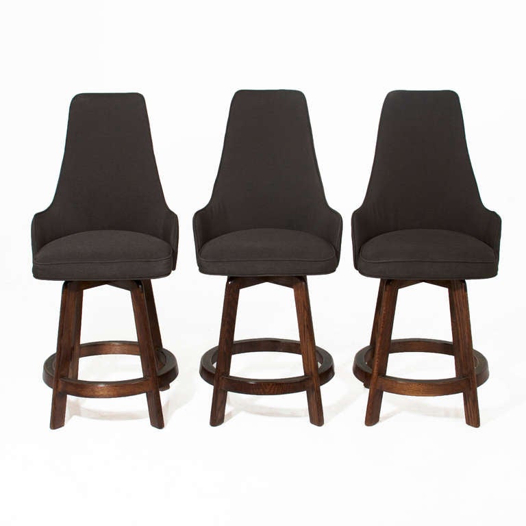 A set of three oak bar stools with swivelling seats with comfortable high backs, upholstered in charcoal fabric. The rounded foot rests have patinated bronze plates to protect the wood. 

Measures: Seat depth 15