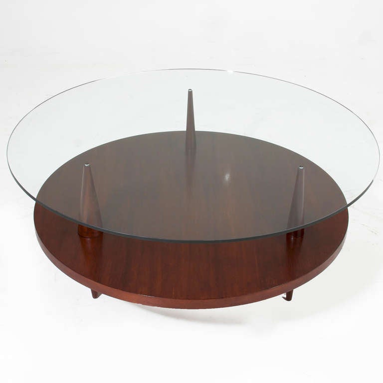 A circular table in solid Brazilian wood with three conical solid wood legs that pierce the round shelf and elegantly support a round glass top. The design is delicate but highly functional and is excellent for storing books or smalls in plain view