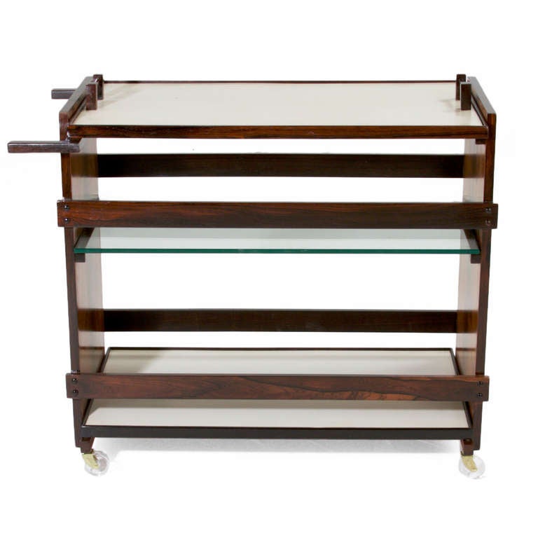A Solid Rosewood rolling bar cart, tea cart, or serving cart from Brazil with three shelves. The top shelf acts as a removable serving tray and is made of white melamine. The middle shelf is made of glass and the bottom shelf is white melamine. All