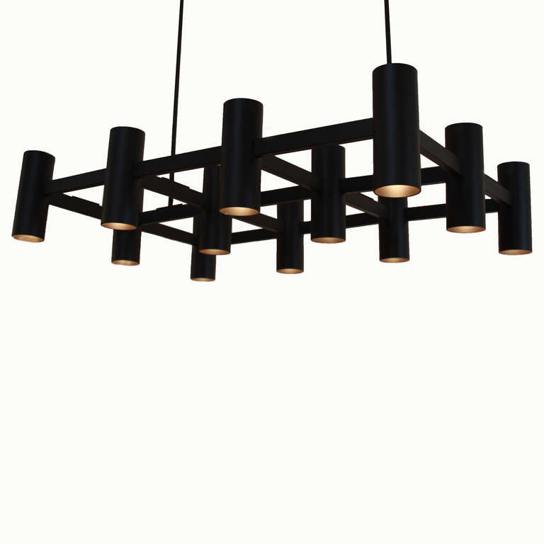 A large chandelier by Thomas Hayes Studio with 12 cylinders on a rectangular support in dark bronze finished steel that appears close to black. The cylinders have both up lights and down lights, and the light makes the inside of the cylinders appear