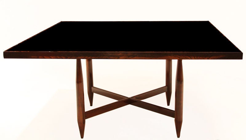 Large dining table with solid rosewood base and dark edge banding around a black reversed painted glass top. Legs are elegantly tapered at the top and bottom and the cross piece adds support.

 