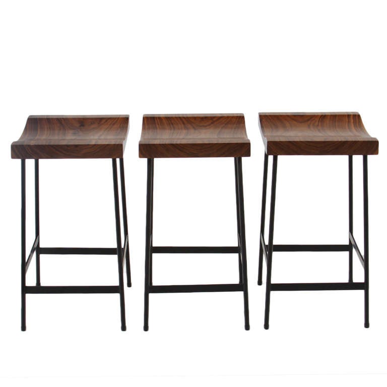 Flat black finished steel bar stools with solid carved figural Walnut seats by Thomas Hayes Studio. 

This item is available for custom order and the lead time is 6-8 weeks; sometimes we are able to complete projects faster, so please contact us