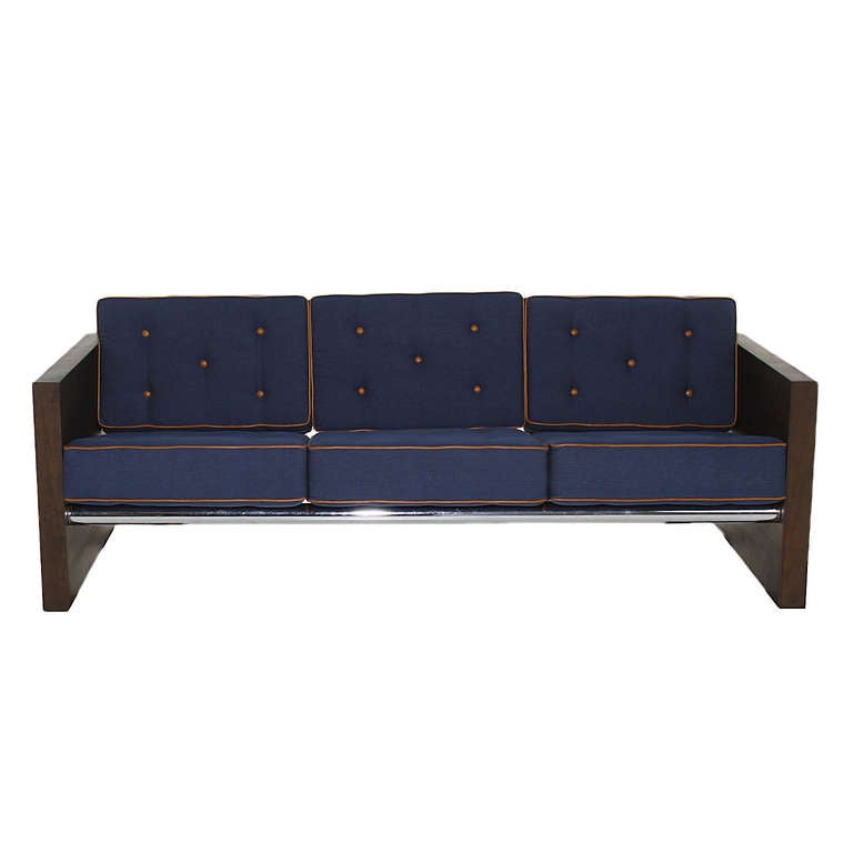 A simple designed sofa with Solid Walnut sides that are in a chocolate oil finish. The seats have been upholstered in a blue denim fabric and have tan leather piping and buttons. The sofa has three tubular chrome bars, two in the back and one below
