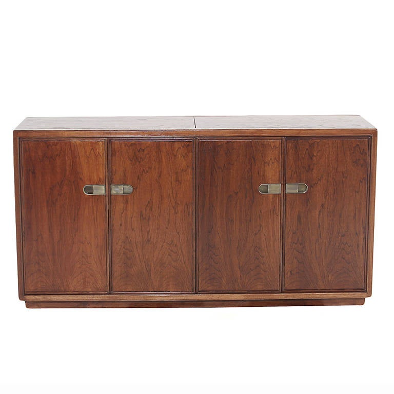 A beautifully made walnut credenza by Drexel with original bronze hardware. The left interior cabinet contains an adjustable shelf, and the right side has four drawers. The top surface has two shelves that extend out to the sides when pulled for