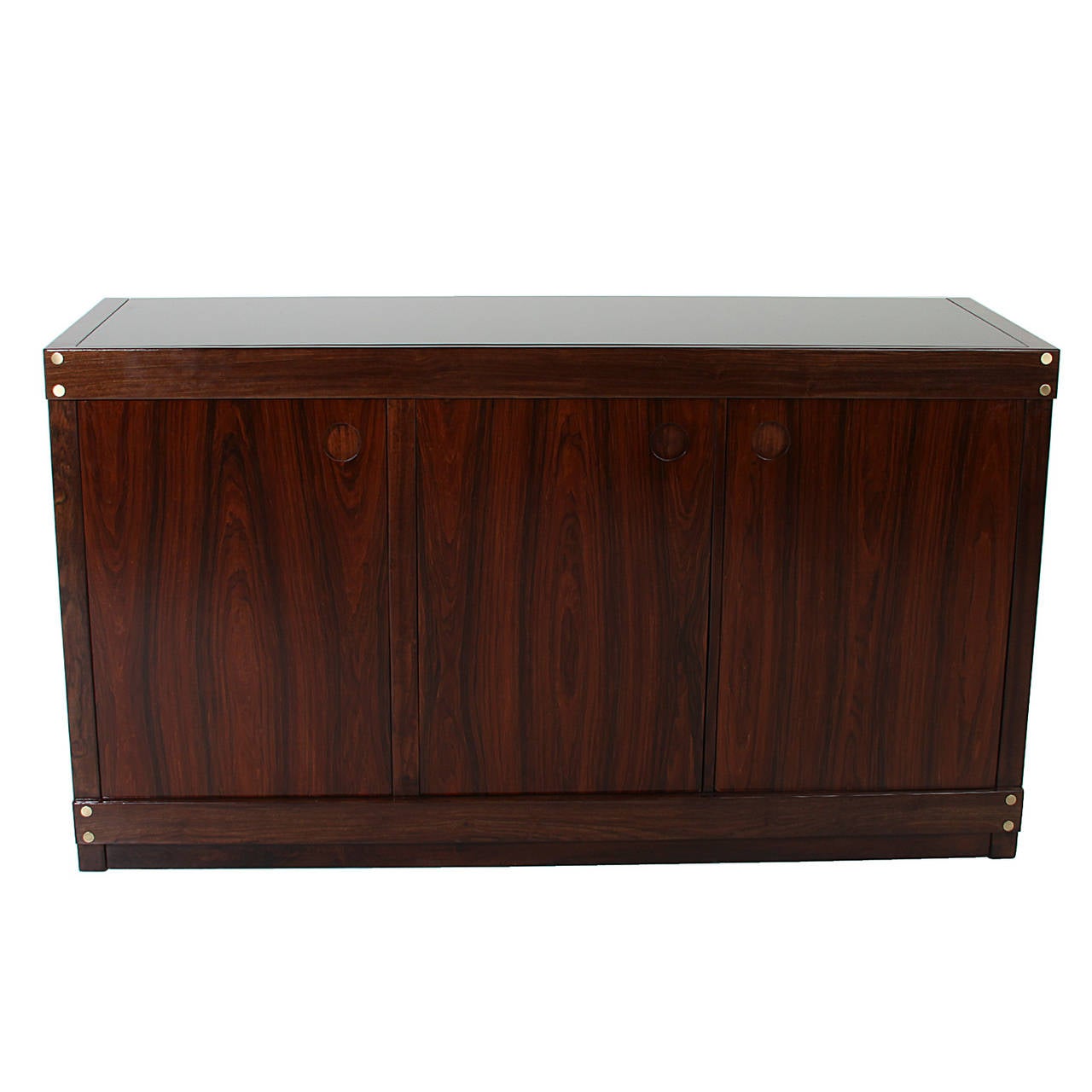 A stunning three-door Rosewood credenza or sideboard from Brazil, designed by Sergio Rodrigues. Featuring new black glass top, inset circular pulls and brass plug details. Newly refinished in Satin Lacquer.

Many pieces are stored in our