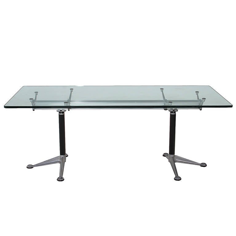 An architectural dining table with a glass top that has rounded edges. The base is made of steel and chrome. The legs have a three legged splay with rounded ends on each foot. The top of the base has a beam made of chrome and steel, there are six