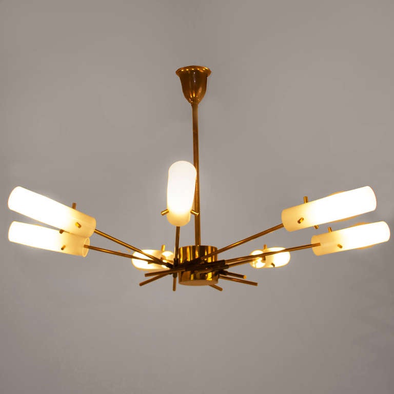 A brass chandelier designed by Arteluce with seven flared spokes with white cylindrical acrylic shades held in place by brass pins. The spokes are affixed to the brass centerpiece with black metal pieces. Retains original patina. Rewired for safety