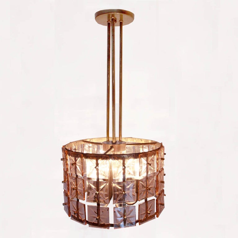 A pair of glass tile and brass chandeliers.  There are three rows of beveled rosé colored glass tiles, secured by bass finials, and arranged altogether in a drum shape.  Above this, three brass bars elegantly lead to the ceiling. These items are