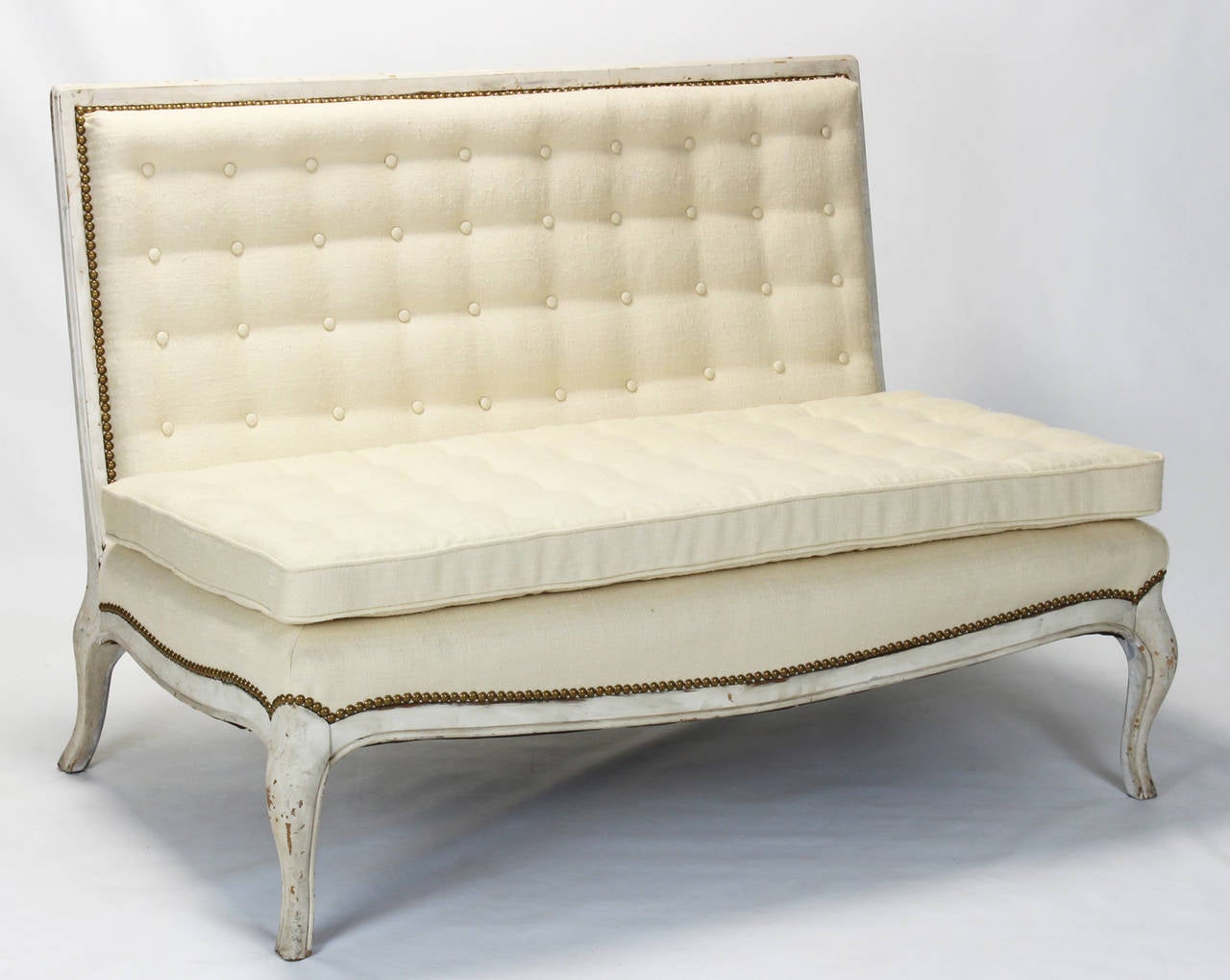 An early 20th century French carved and painted wood settee or bench button upholstered in cream colored linen fabric and accented with brass nailheads.