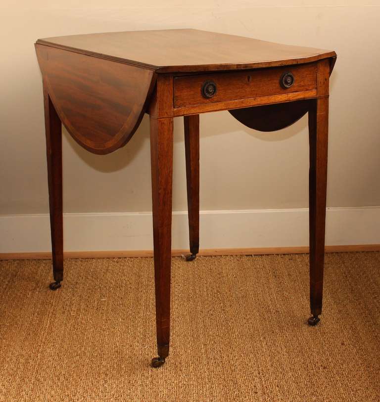 A late 18th century English small scale Pembroke table in mellow sun faded mahogany with satin wood accents and original brass pulls resting on delicate tapering legs terminating in brass casters.