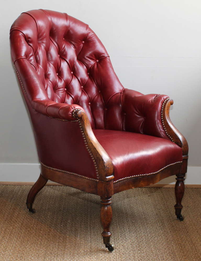 An early 19th century English barrel style buttoned library chair covered in deep red leather and accented with brass nail heads resting on turned mahogany legs terminating in brass casters.