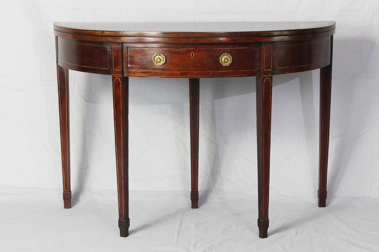 An early 19th century Hepplewhite mahogany demilune card table with satinwood inlay and rosewood banded top. The table is supported by five square tapering legs, the two outside rear being gate legs. The front apron displays a single operating