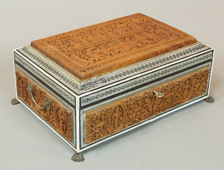 An exceptionally large Anglo Indian sewing box. The top and all four sides are heavily and exquisitely carved, surrounded by ivory and ebony accents resting on brass paw feet. The interior of the box displays many lidded compartments.