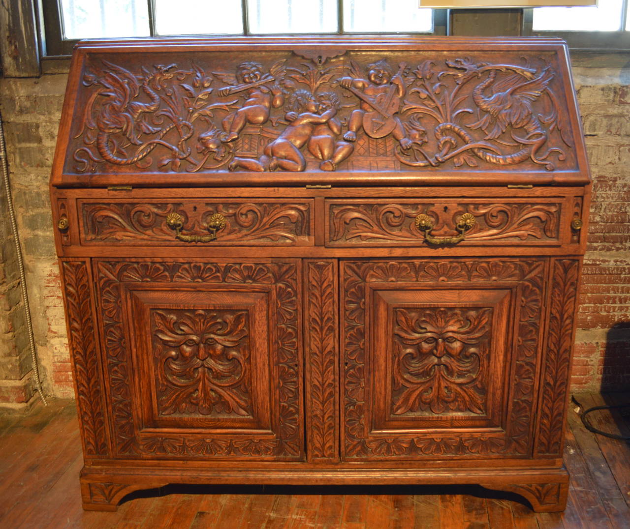 Unusually carved Italian Renaissance style desk, central fall panel of band of putti playing string instruments, fitted interior, prospect door with carved green man, slant fall front, lower section with two drawers over two-paneled doors with green