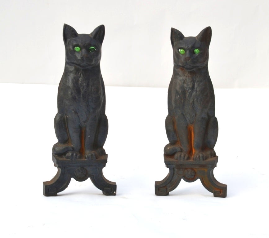 A pair of vintage cast iron andirons in the shape of seated black cats. Each has green glass eyes and an aged, rust accented patina