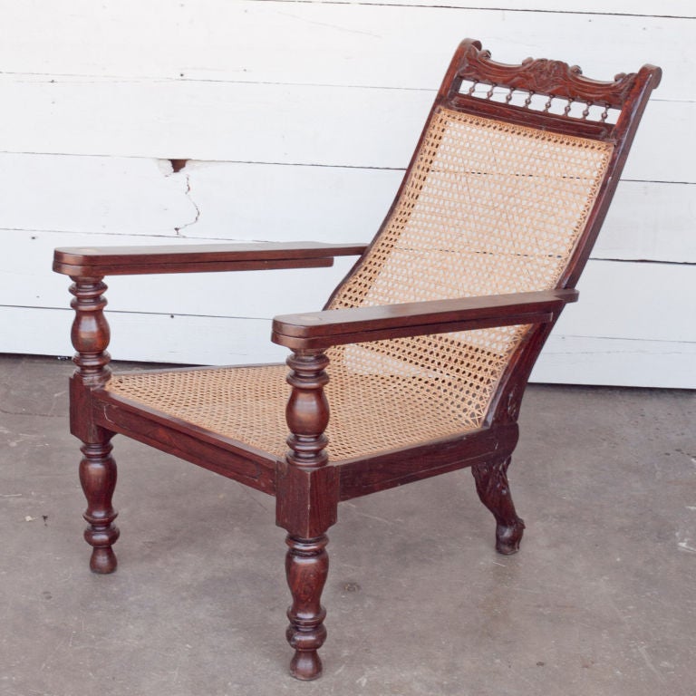 Anglo-Indian solid rosewood plantation chair with carved details on back and caned seat. Arms extend out as foot rests. The back feet are carved in the shape of a stylized animal head.