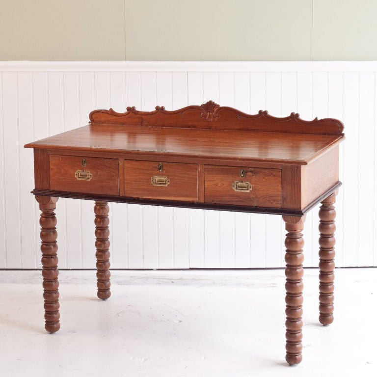 Anglo-Indian solid teak desk resting on bobbin style turned legs. Three drawers with inset brass handles. Solid rosewood trim along bottom edge of desk. Back of table has carved and scalloped top edge.