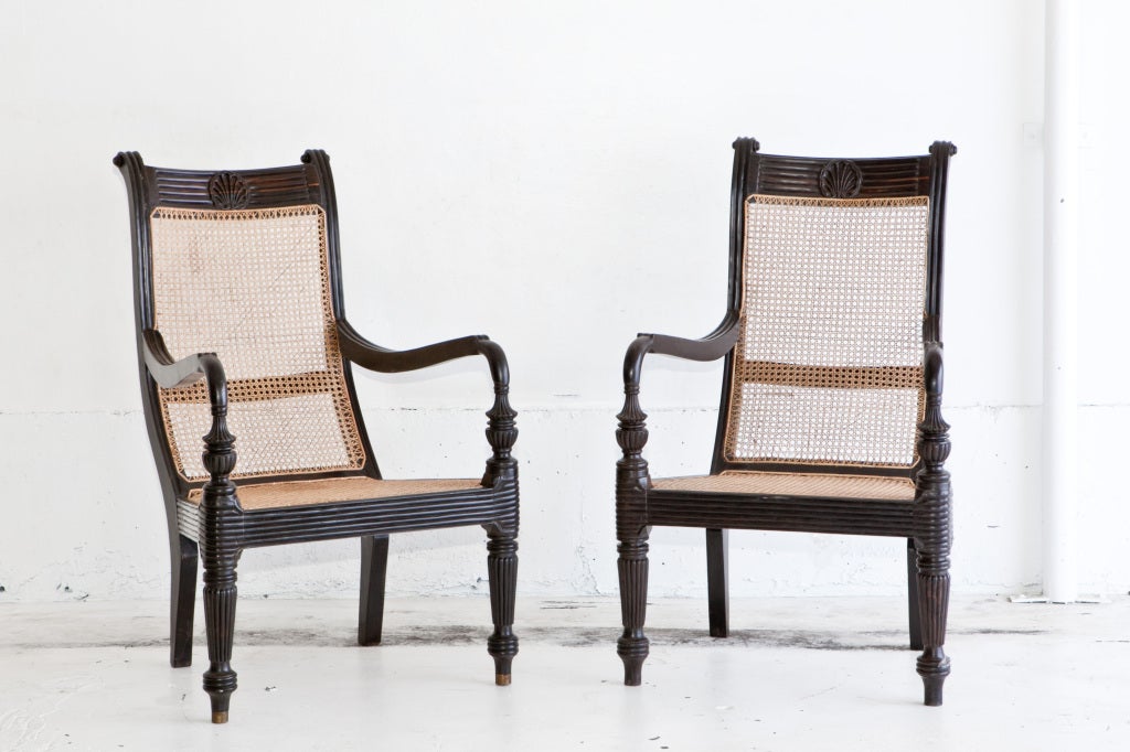 British colonial Grandfather chair of solid hand-carved ebony with caned seat. Seat aprons and legs have carved fluting, back has scallop design. One chair has brass rings on the bottom feet, the other does not otherwise they are identical. Chairs