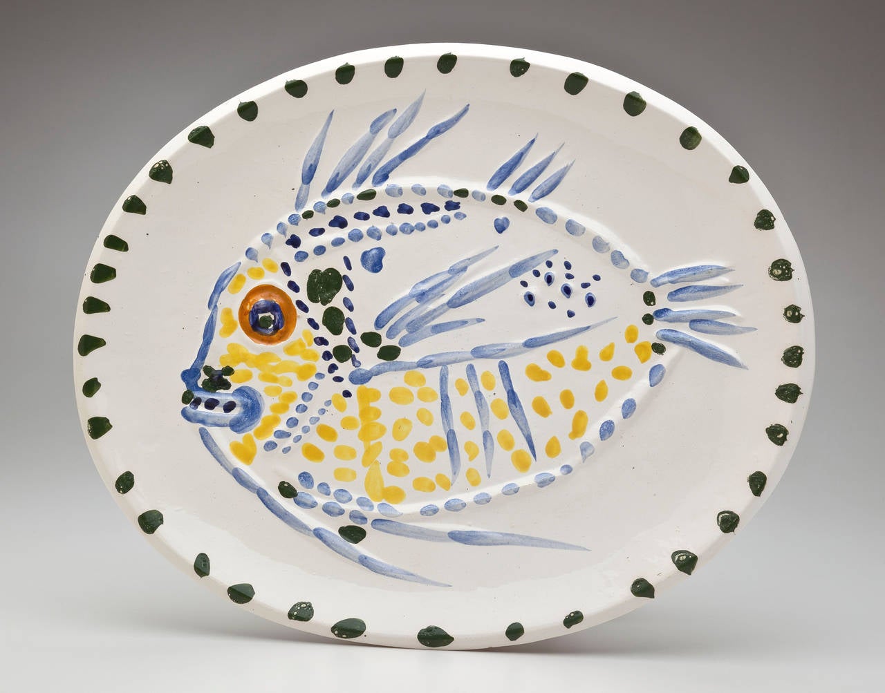 A large ceramic platter with a Fish Design by Pablo Picasso (1881-1973). The spotted yellow and blue fish with an orange eye dominates the glazed oval earthenware dish. The white enamel surface is covered with dots of yellow, green, orange and blue