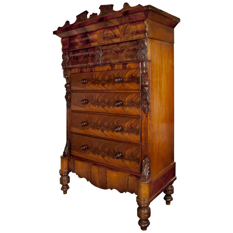 Monumental carved mahogany and flame mahogany veneer chest of drawers, bookmatched veneer on the curved drawer fronts, carved sunflower motifs flank the broken pediment top and cascade down the pilasters, scalloped apron, bulbous turned feet, turned