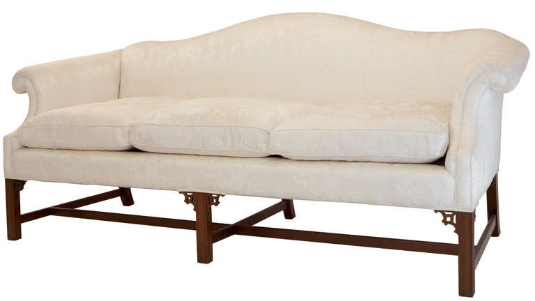 Nicely shaped Chippendale style camelback sofa, scooped back scrolled arms, mahogany base with straight legs and stretchers, Chinese fretwork brackets top the front legs, covered in a fine cream colored damask with Oriental scenes, three seat